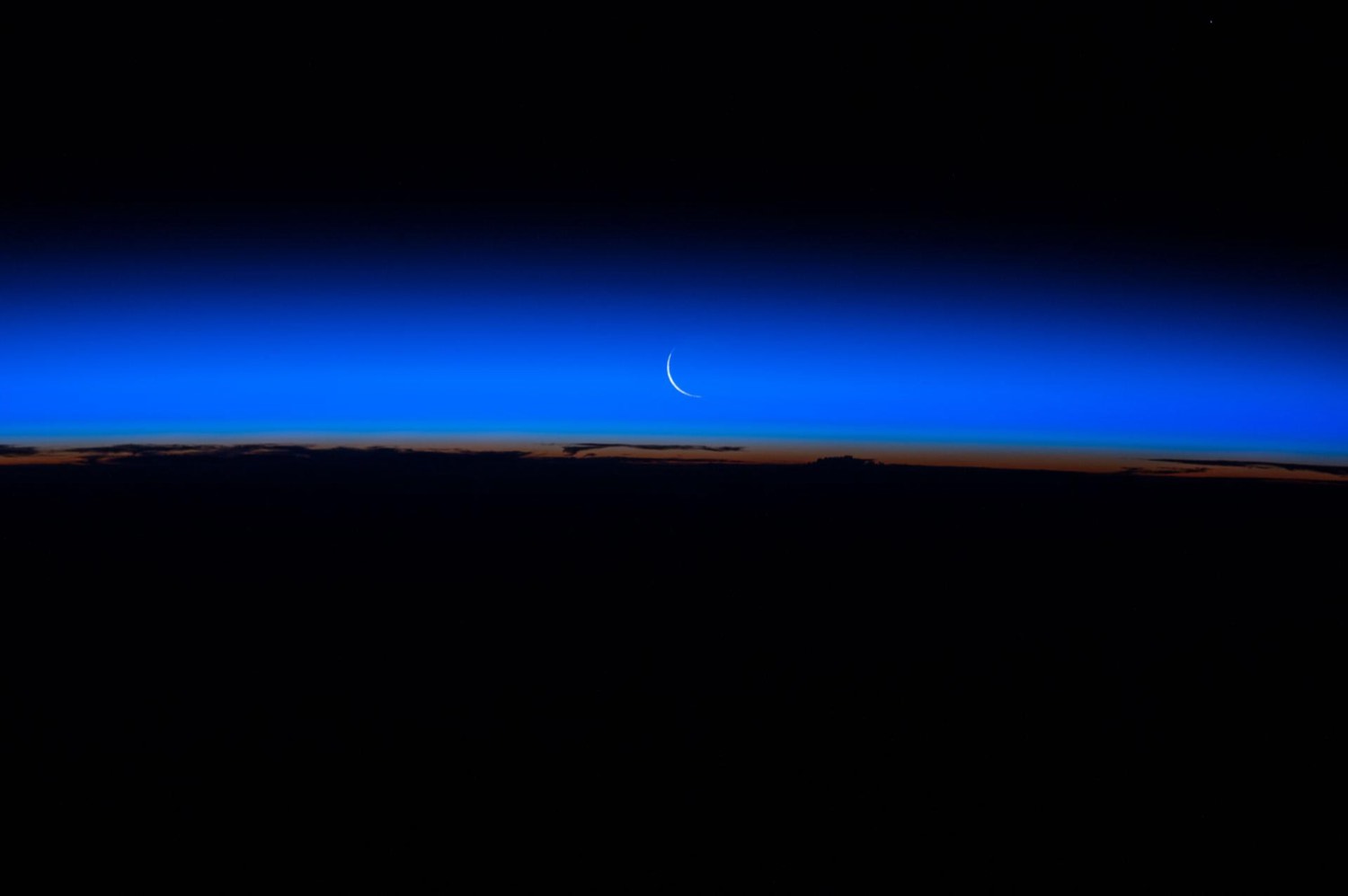 A thin crescent Moon above the layered blue and orange glow of Earth's horizon