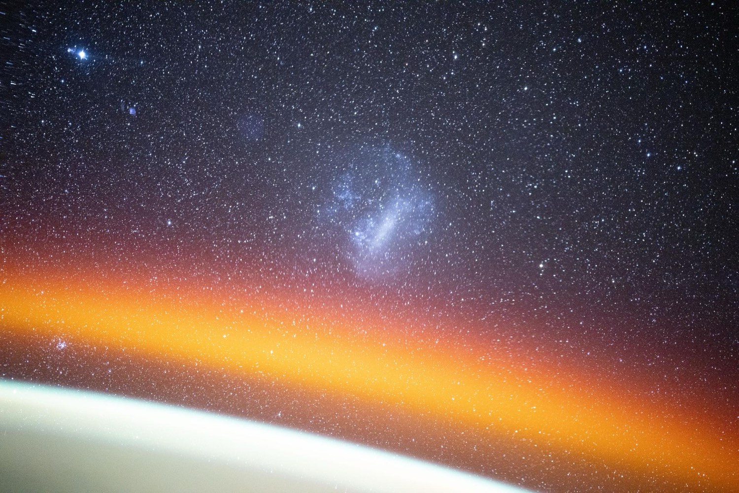 The Large Magellanic Cloud as a soft glowing patch among stars above the thin orange glow of Earth's atmosphere