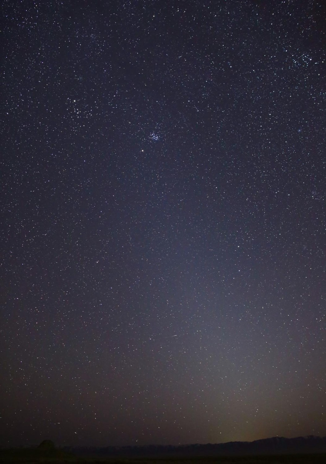 A tall, faint cone of light rising from a dark desert horizon into a starry sky, with the Pleiades near the top